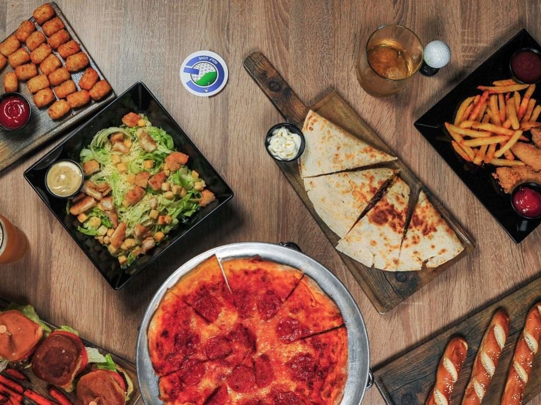 A spread of Golf Cove food including a pepperoni pizza, quesadilla, Caesar salad, tots, sliders, tenders with fries and pretzels