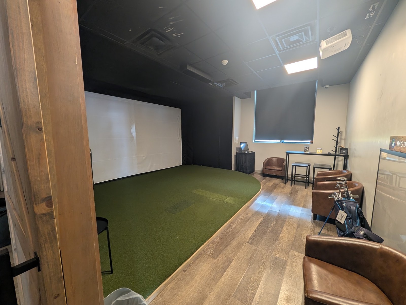A Golf Cove simulator bay with a hitting mat, screen, leather chairs and a rental golf bag