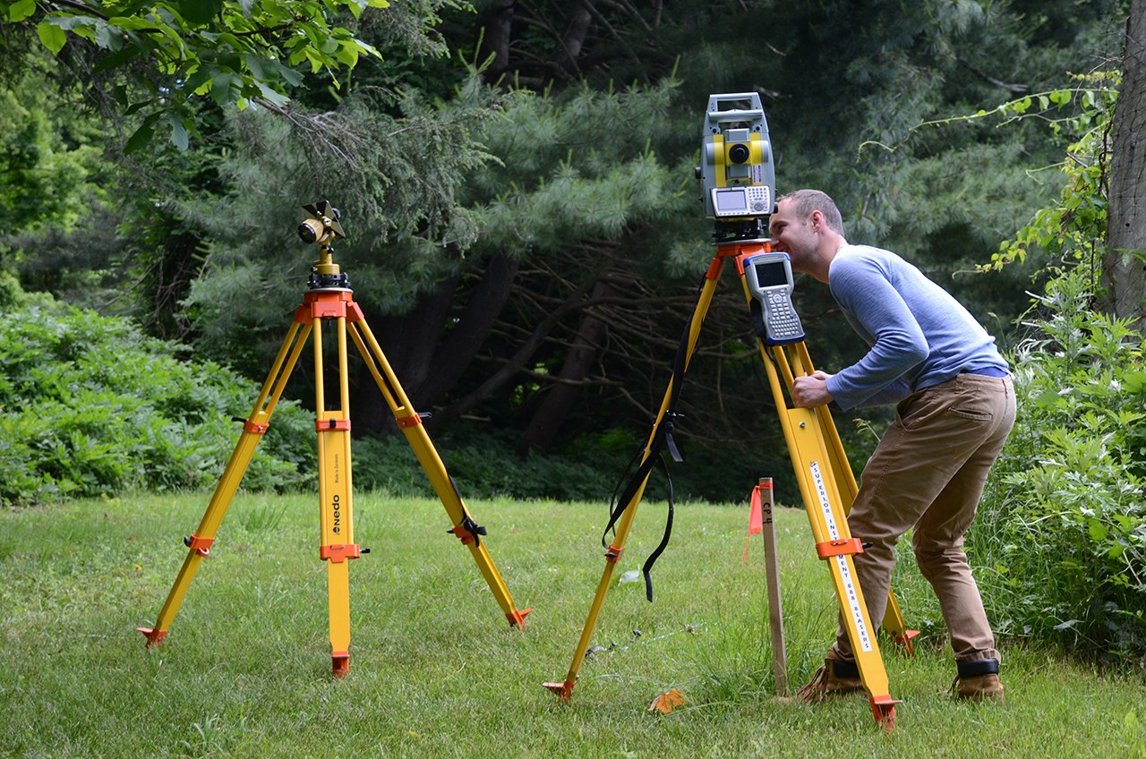 A man sets up survey equipment on yellow tripods in a grassy clearing at Wah Wah Taysee
